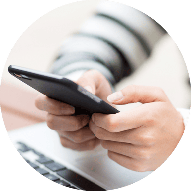 Gros plan sur des mains tenant un téléphone mobile, avec un ordinateur portable flou en arrière-plan.